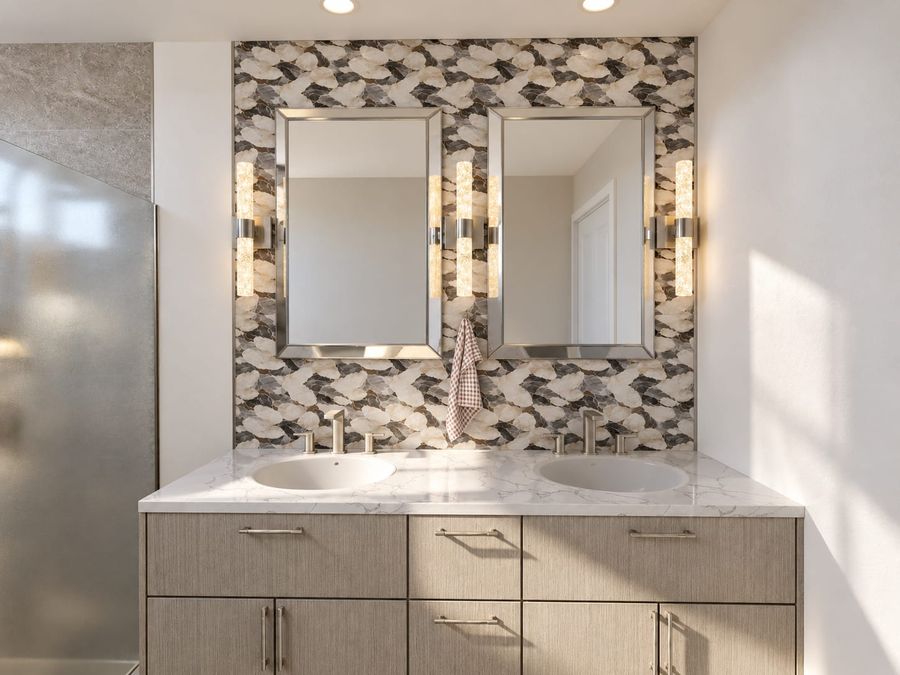 3D rendering of a bathroom with a gray wood double vanity, a marble-look counter, two framed mirrors with crystal sconces, and a floor-to-ceiling cream, gray, and brown pebble mosaic wall