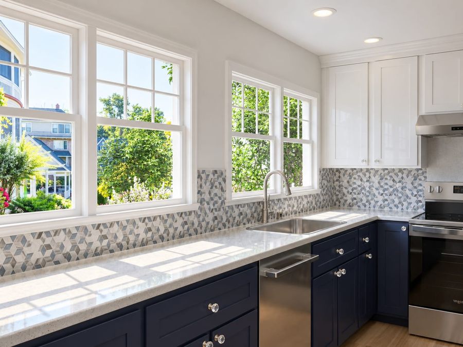 3D rendering of the same kitchen's sink wall: two sets of white double-hung windows, the geometric mosaic backsplash, quartz counters, and navy cabinets with crystal knobs